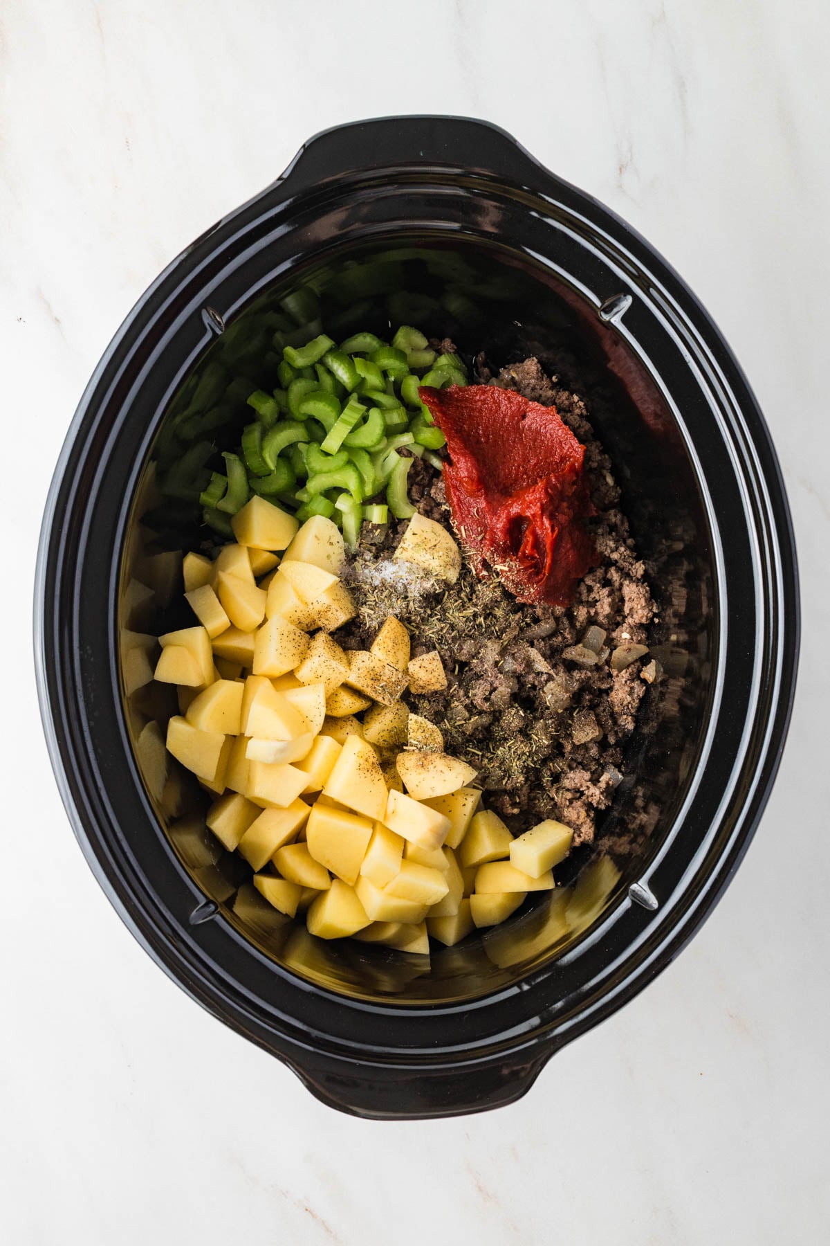 Slow cooker filled with chopped potatoes, ground beef, celery, tomato paste, and seasonings, ready to be cooked.