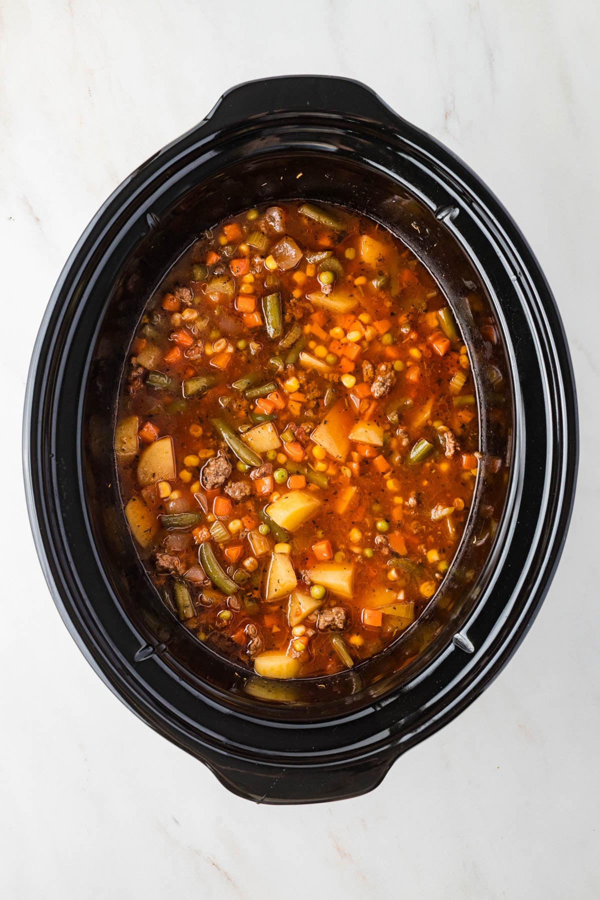 A slow cooker filled with hamburger soup containing potatoes, carrots, peas, corn, green beans, and broth on a white surface.