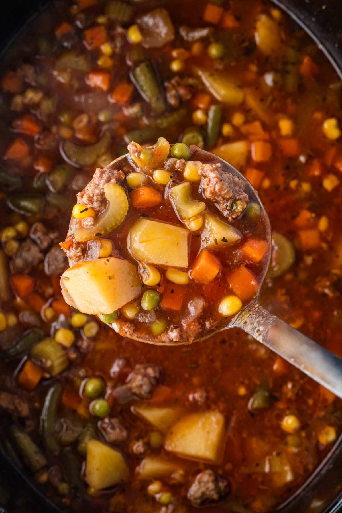Ladle holding hamburger soup with potatoes, carrots, peas, corn, green beans, and ground beef in a tomato-based broth.