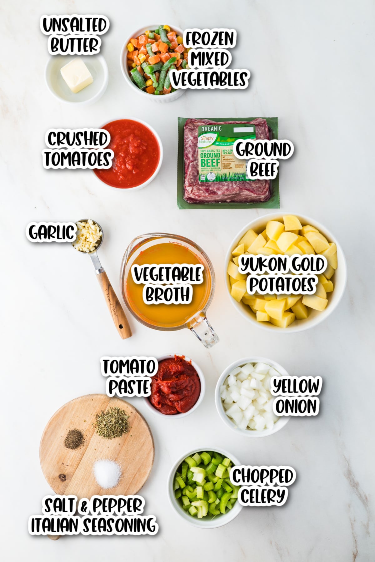 Various ingredients for a soup or stew are arranged on a white surface, including ground beef, potatoes, broth, vegetables, tomato paste, garlic, onion, celery, butter, and seasonings.