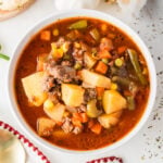 A bowl of hamburger soup with potatoes, carrots, corn, green beans, and ground beef in a red broth sits on a white surface.