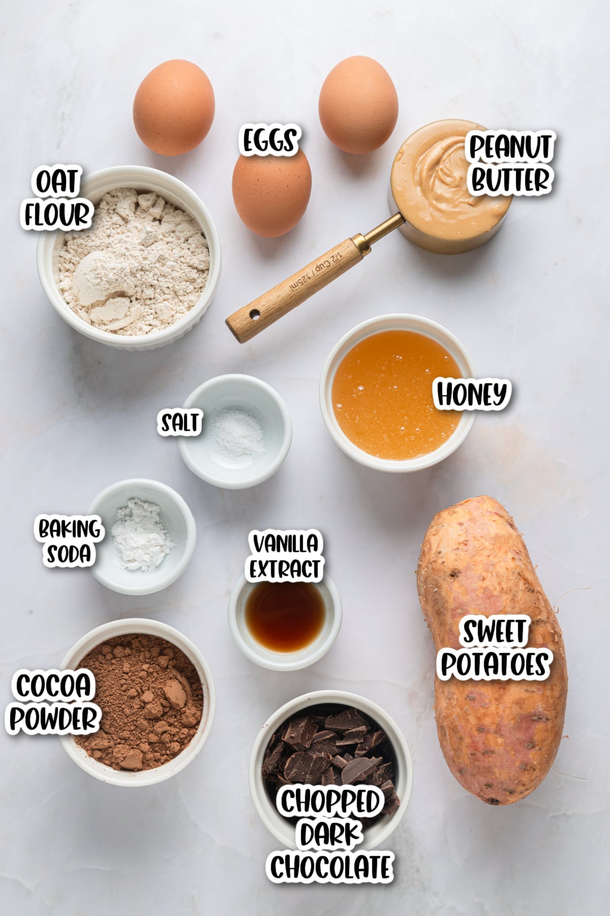 Top-down view of baking ingredients in bowls, including eggs, peanut butter, oat flour, honey, salt, baking soda, vanilla extract, cocoa powder, chopped dark chocolate, and a sweet potato.