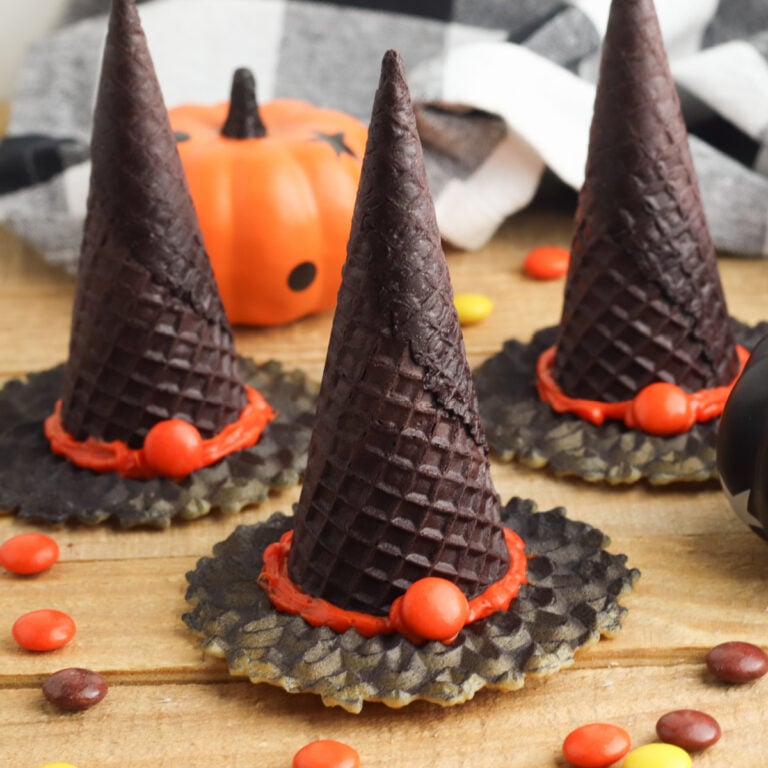 Three edible witch hats made from chocolate wafer cones and cookies, decorated with red icing and candy, are displayed on a wooden surface with Halloween props in the background.