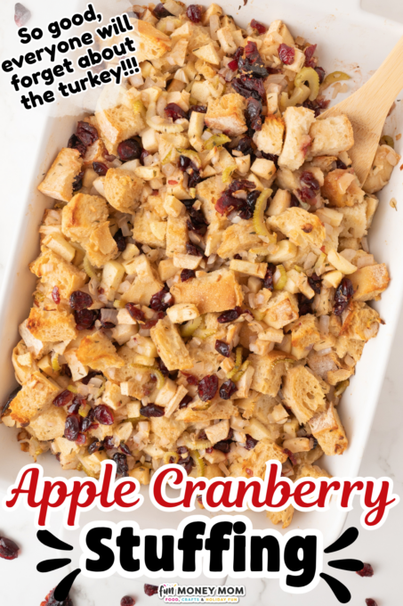 A casserole dish filled with apple cranberry stuffing, containing bread cubes, cranberries, and apple pieces, with a wooden spoon on the side.