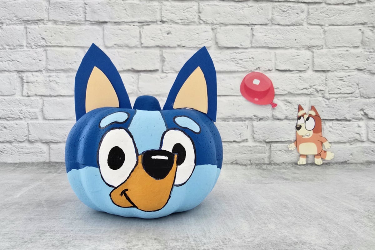 A pumpkin painted to look like Bluey sits in front of a white brick wall, with a small figure holding a red balloon in the background.