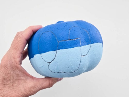 A hand holds a blue pumpkin with a mouse face outline drawn on it, against a plain white background.