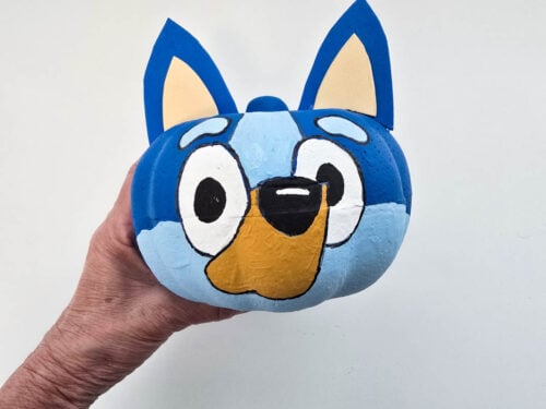 A hand holds a blue pumpkin painted with the face of a cartoon dog, featuring large eyes, blue ears, and a tan snout.