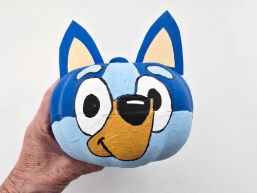A hand holds a pumpkin painted to resemble a blue cartoon dog with large eyes, blue ears, and a yellow snout, against a plain white background.