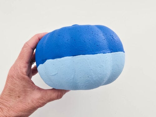 A hand holds a foam craft pumpkin, painted dark blue on the top half and light blue on the bottom half, against a plain white background.