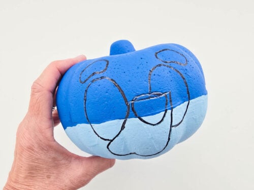 A hand holding a pumpkin painted blue with a cartoon face outlined in black on its surface against a plain white background.