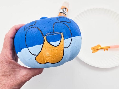 A hand holds a small pumpkin painted blue with a cartoon face. A paintbrush with light brown paint and a bottle of paint are nearby on a white surface.