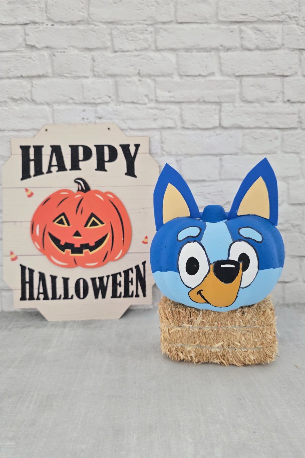 A painted pumpkin resembling a blue cartoon dog sits on hay beside a “Happy Halloween” sign with a jack-o’-lantern, against a white brick wall.