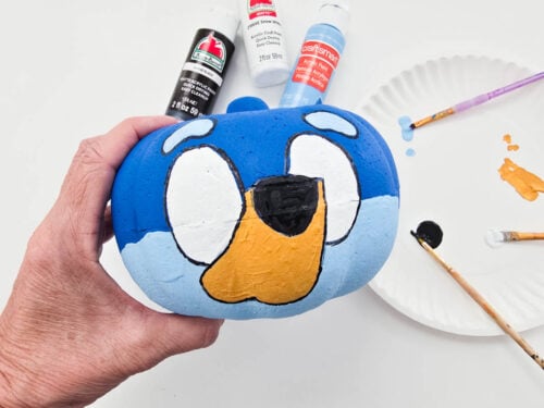 A hand holds a painted pumpkin featuring a cartoon dog’s face. Paint bottles, a paper plate, and paintbrushes are visible in the background.