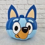 A pumpkin painted to resemble a blue cartoon dog with large eyes, a tan snout, and pointy blue ears, set against a white brick background.