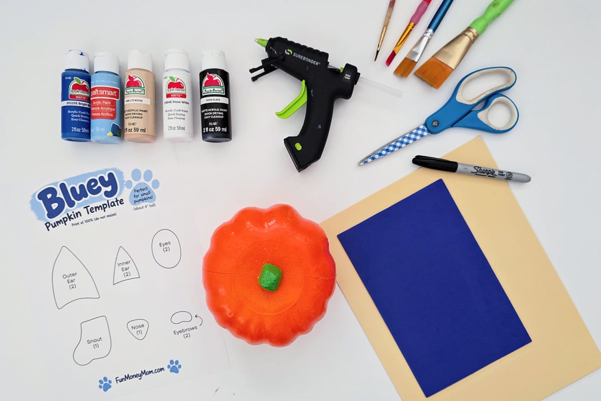 Pumpkin craft supplies arranged on a white surface, including paint, brushes, glue gun, scissors, Sharpie, colored paper, a pumpkin, and a Bluey pumpkin template.
