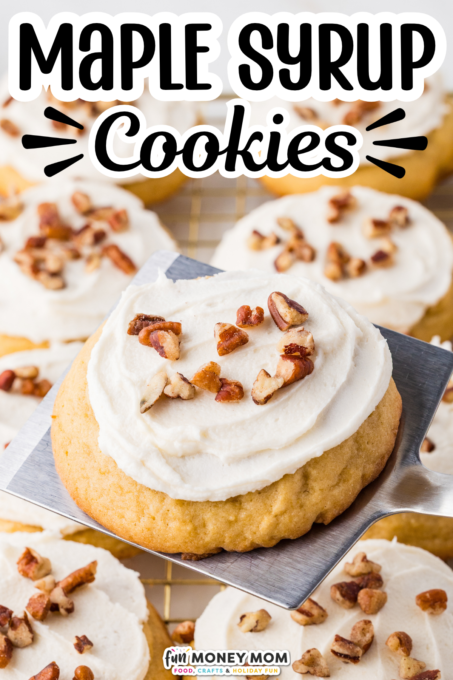 A metal spatula lifts a frosted maple syrup cookie topped with pecans, with more cookies in the background.