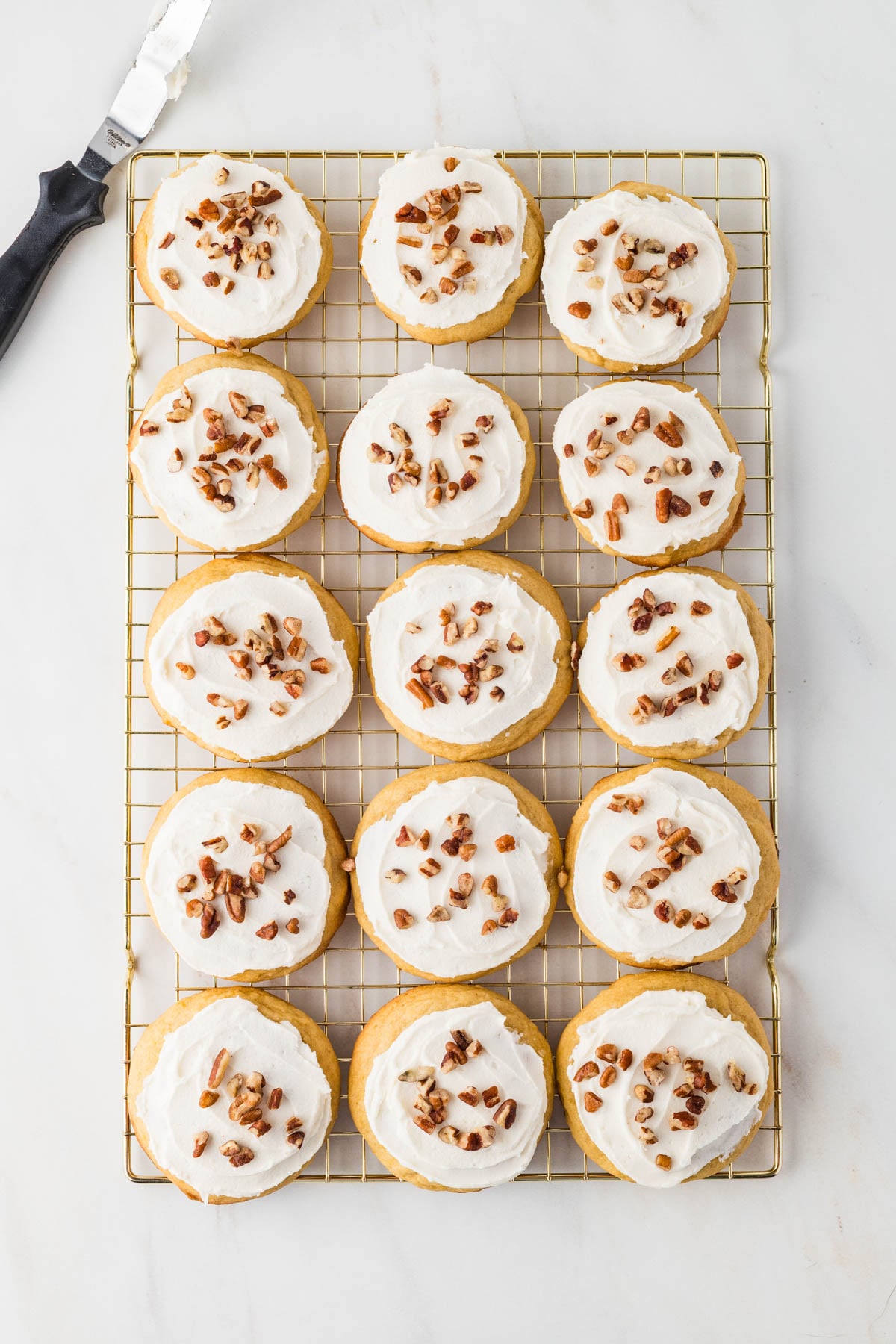 Fifteen round cookies topped with white frosting and chopped pecans, arranged in a grid on a wire cooling rack.