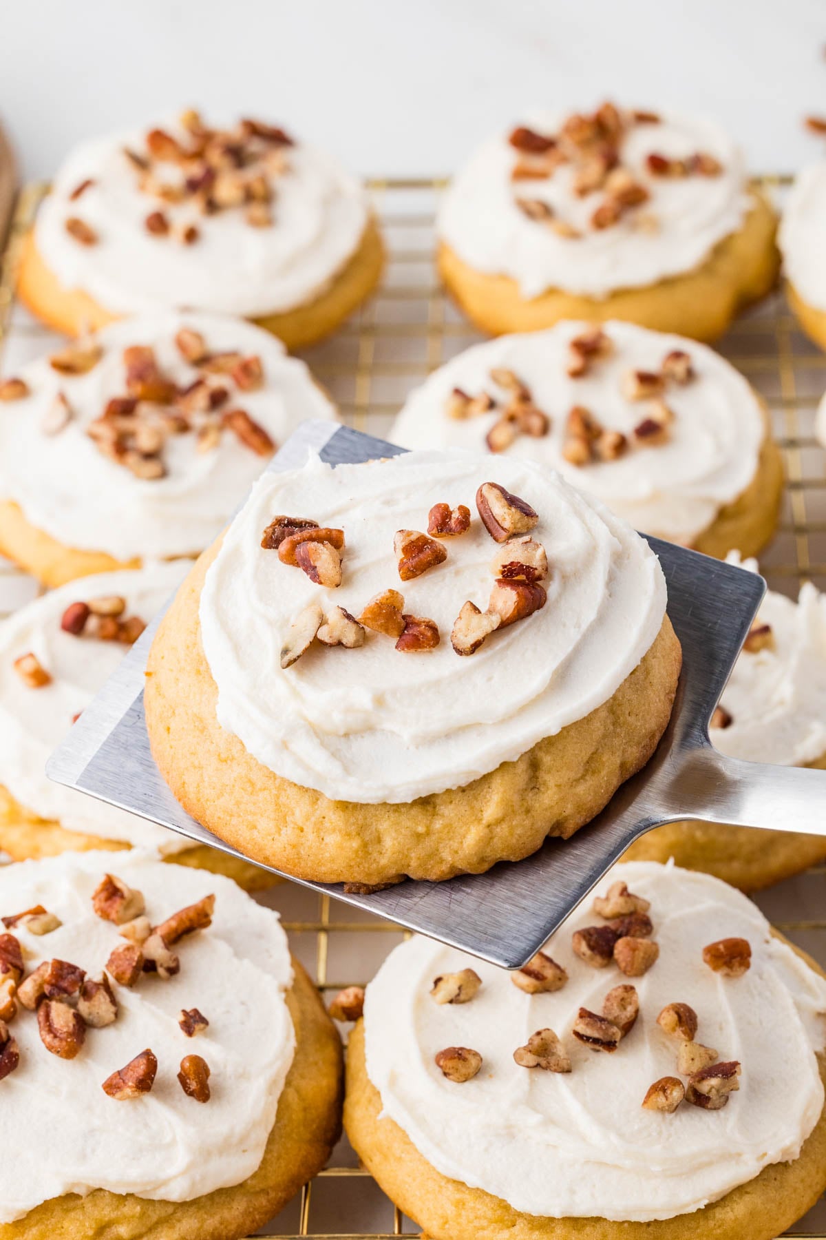 A spatula holds a frosted cookie topped with chopped pecans above a cooling rack filled with similar cookies.