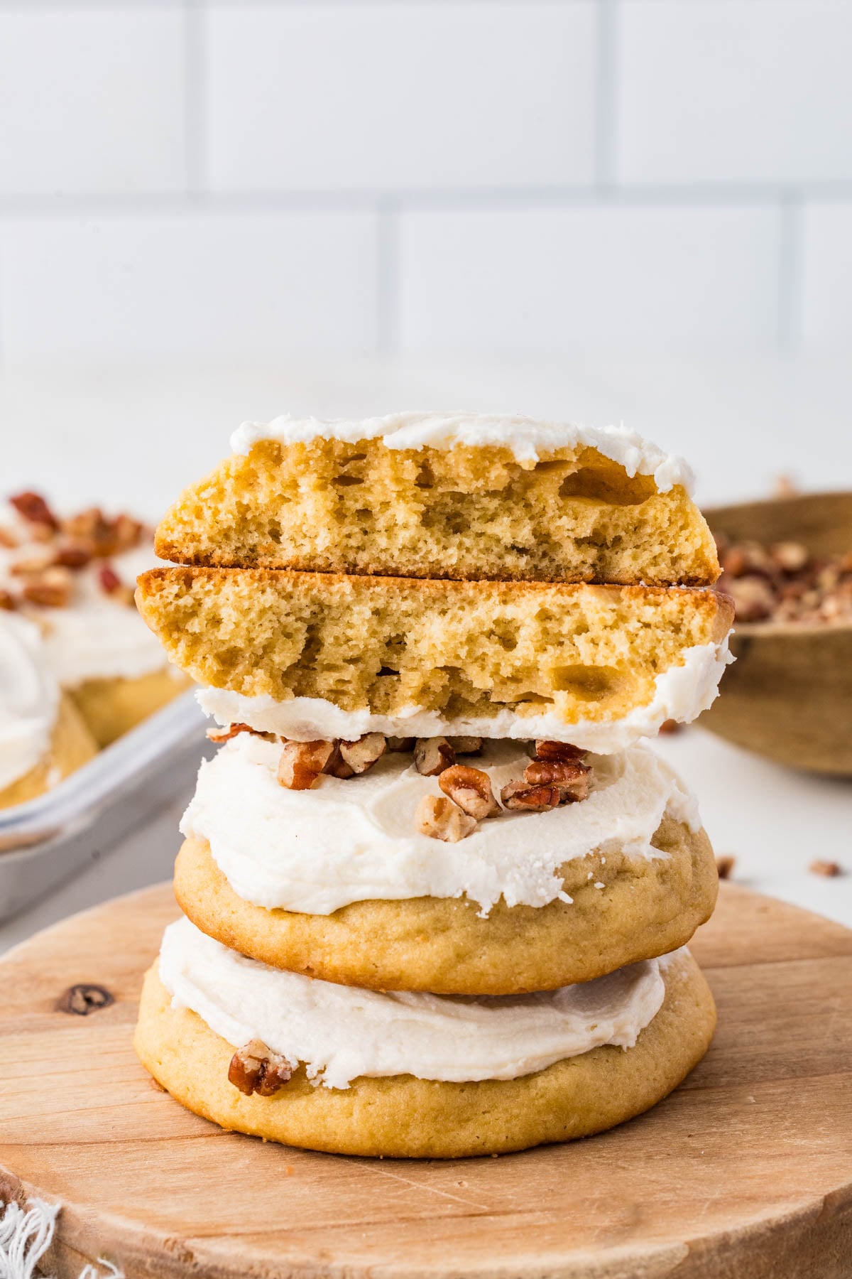 A stack of three frosted cookies topped with pecans on a wooden board, with the top cookie broken in half to show the inside.