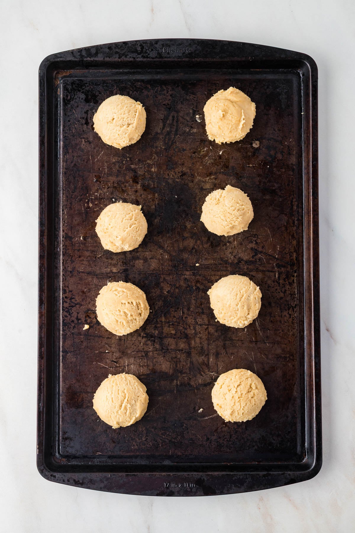 Eight scoops of raw cookie dough arranged in two rows on a dark baking sheet.