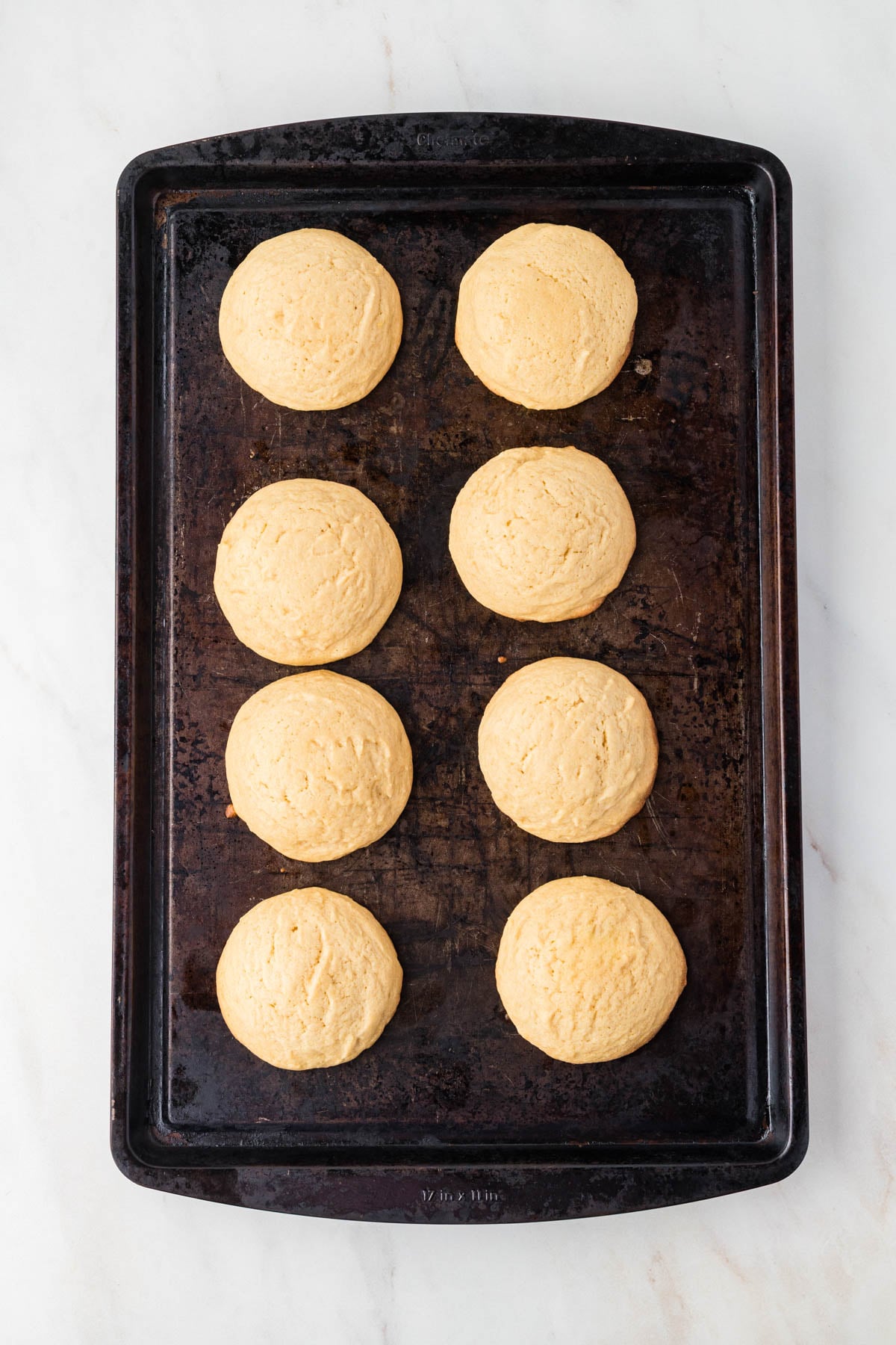 Eight round, baked cookies arranged in two columns on a dark metal baking sheet.