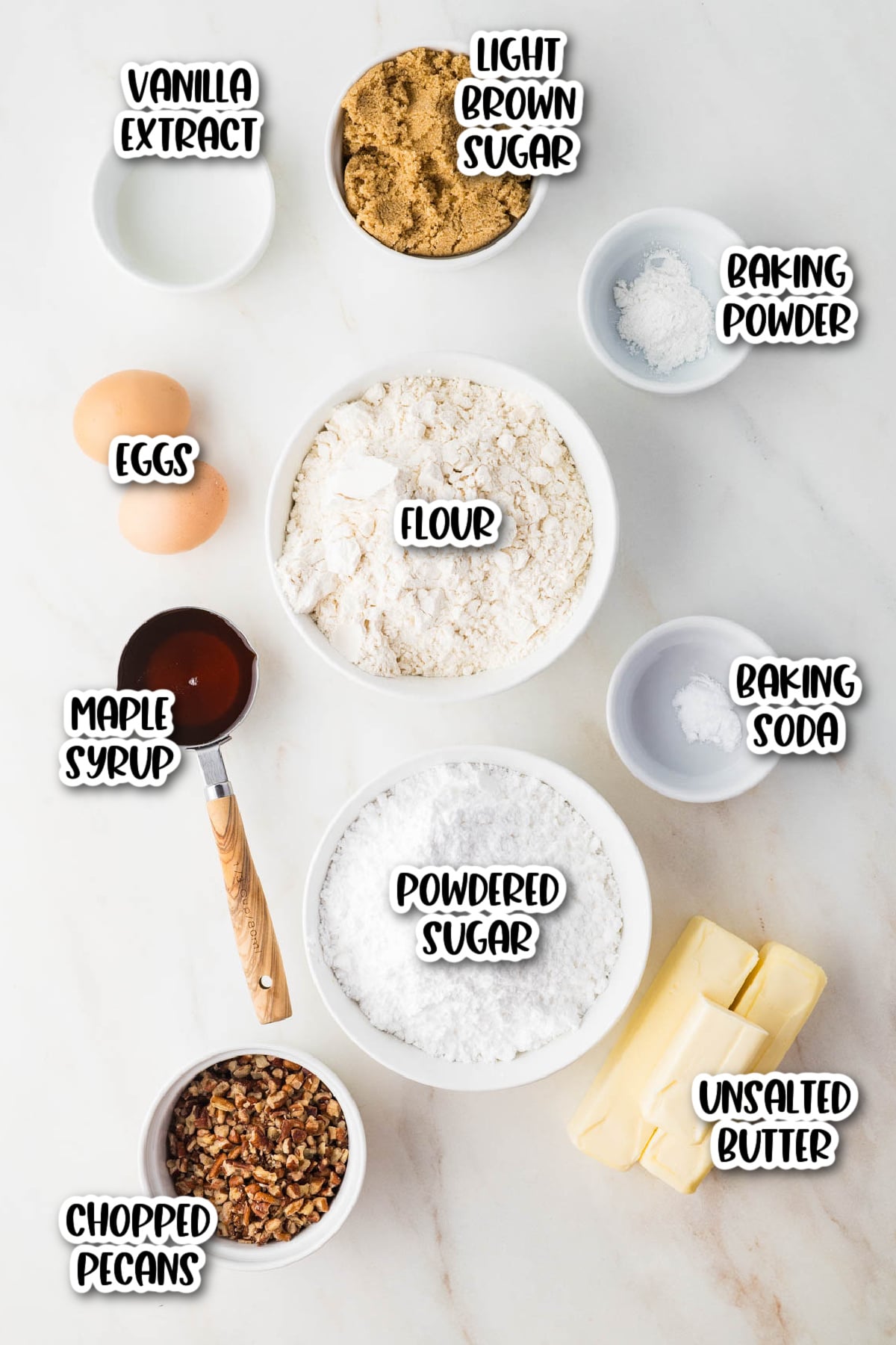 Top-down view of baking ingredients labeled: vanilla extract, brown sugar, baking powder, eggs, flour, maple syrup, baking soda, powdered sugar, chopped pecans, and unsalted butter.