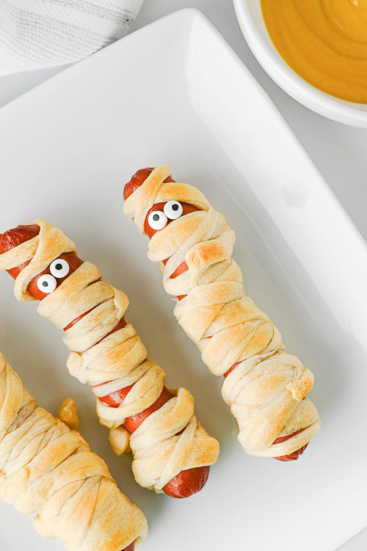 Hot dogs wrapped in dough strips to look like mummies with candy eyes, served with a side of mustard.