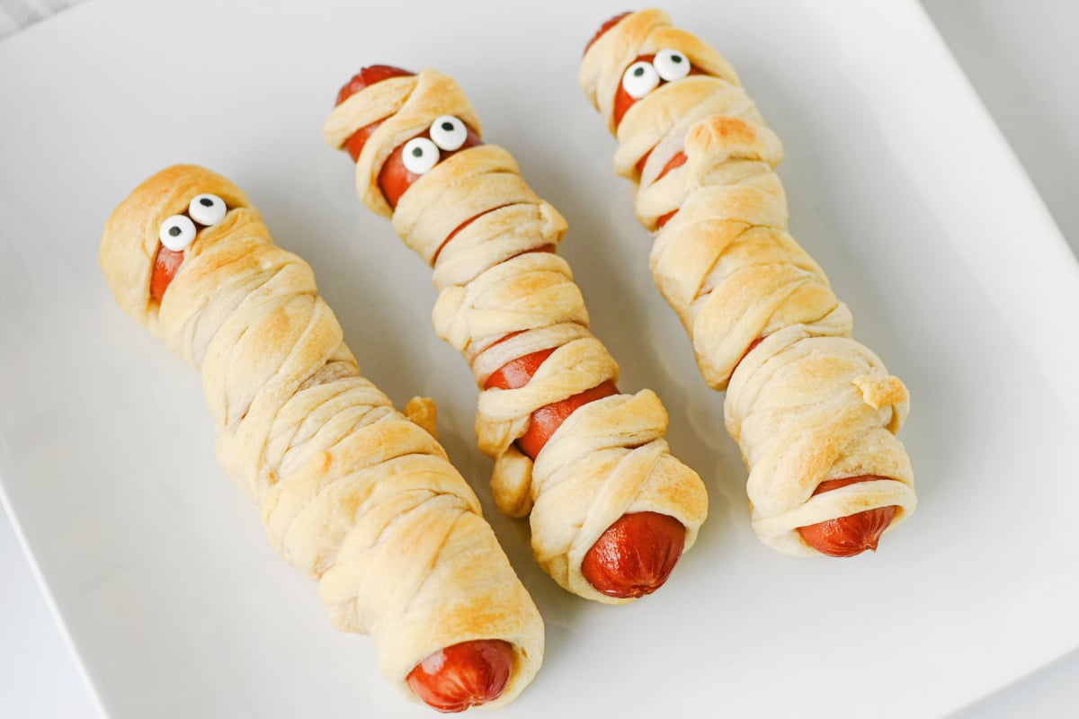 Three hot dogs wrapped in dough strips to look like mummies, each with two edible candy eyes.