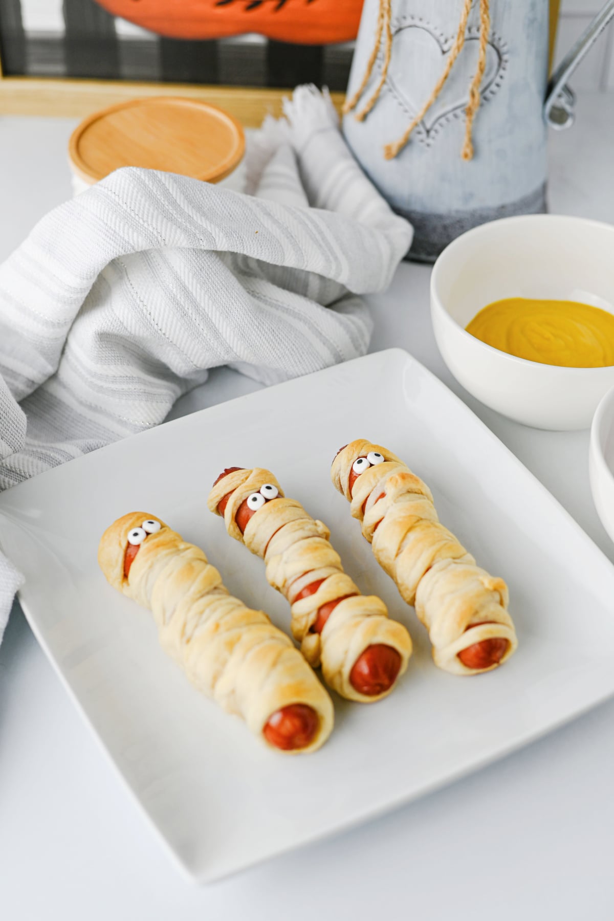 Three hot dogs wrapped in dough to look like mummies with candy eyes, served on a white square plate.