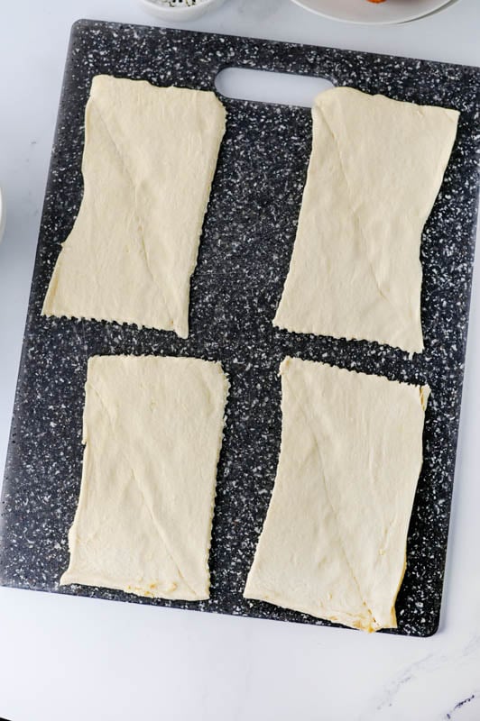 Four rectangular pieces of raw crescent roll dough laid out on a dark cutting board.