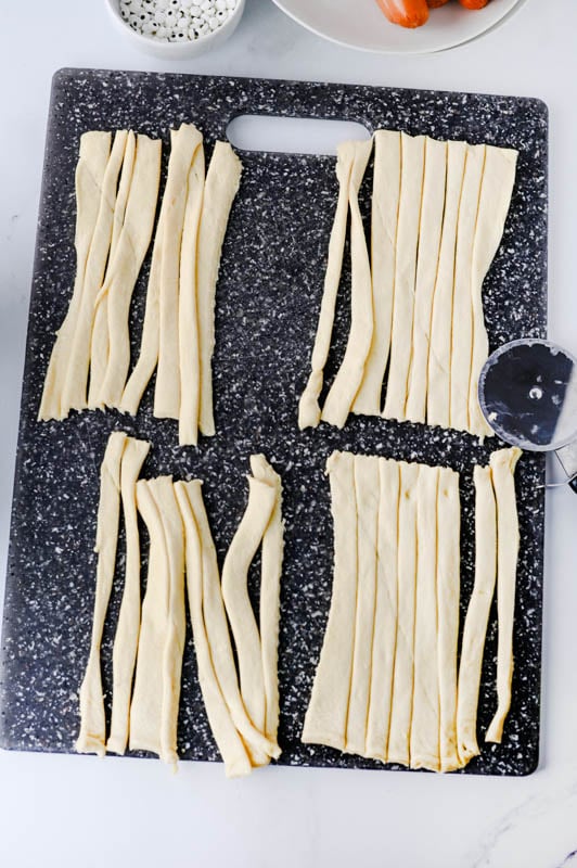 Strips of raw dough arranged in four rectangular groupings on a black cutting board, with a pizza cutter to the side.