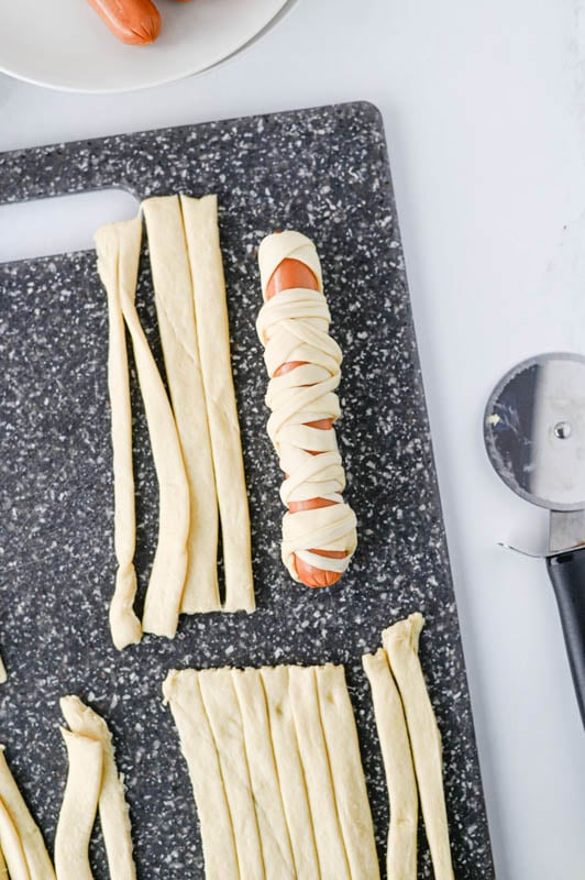 A hot dog being wrapped in thin strips of dough on a cutting board, with a pizza cutter nearby.