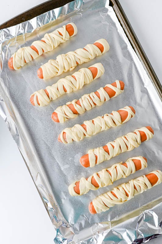 Eight hot dogs wrapped in strips of dough to resemble mummies, arranged on a foil-lined baking sheet.
