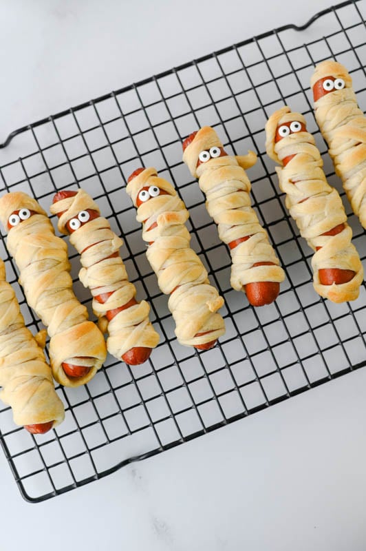 Hot dogs wrapped in strips of dough to look like mummies, complete with candy eyes, resting on a wire cooling rack.