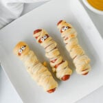Three hot dogs wrapped in dough strips to resemble mummies with candy eyes, served on a square white plate with a side of mustard.