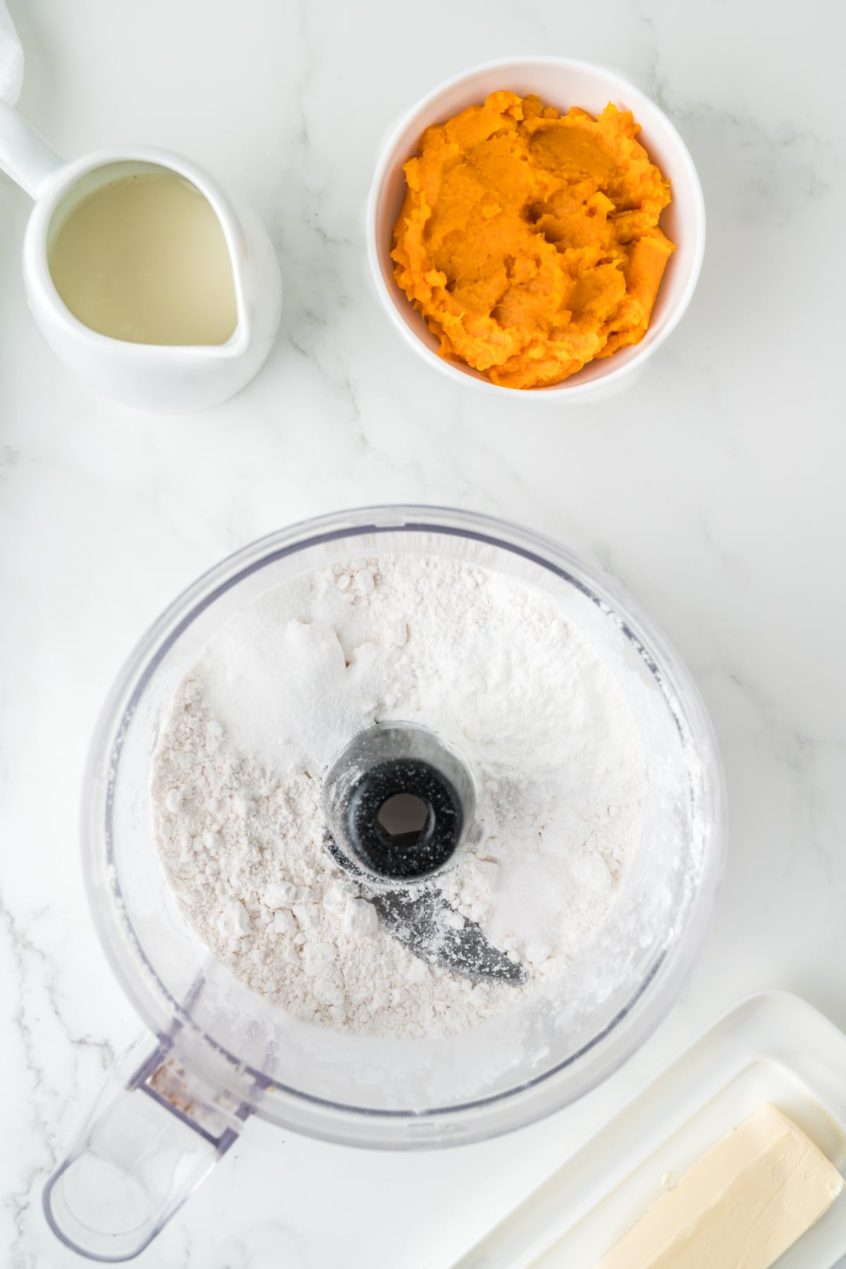 A food processor containing flour, a small bowl of mashed sweet potato, a pitcher of milk, and a stick of butter on a white surface.