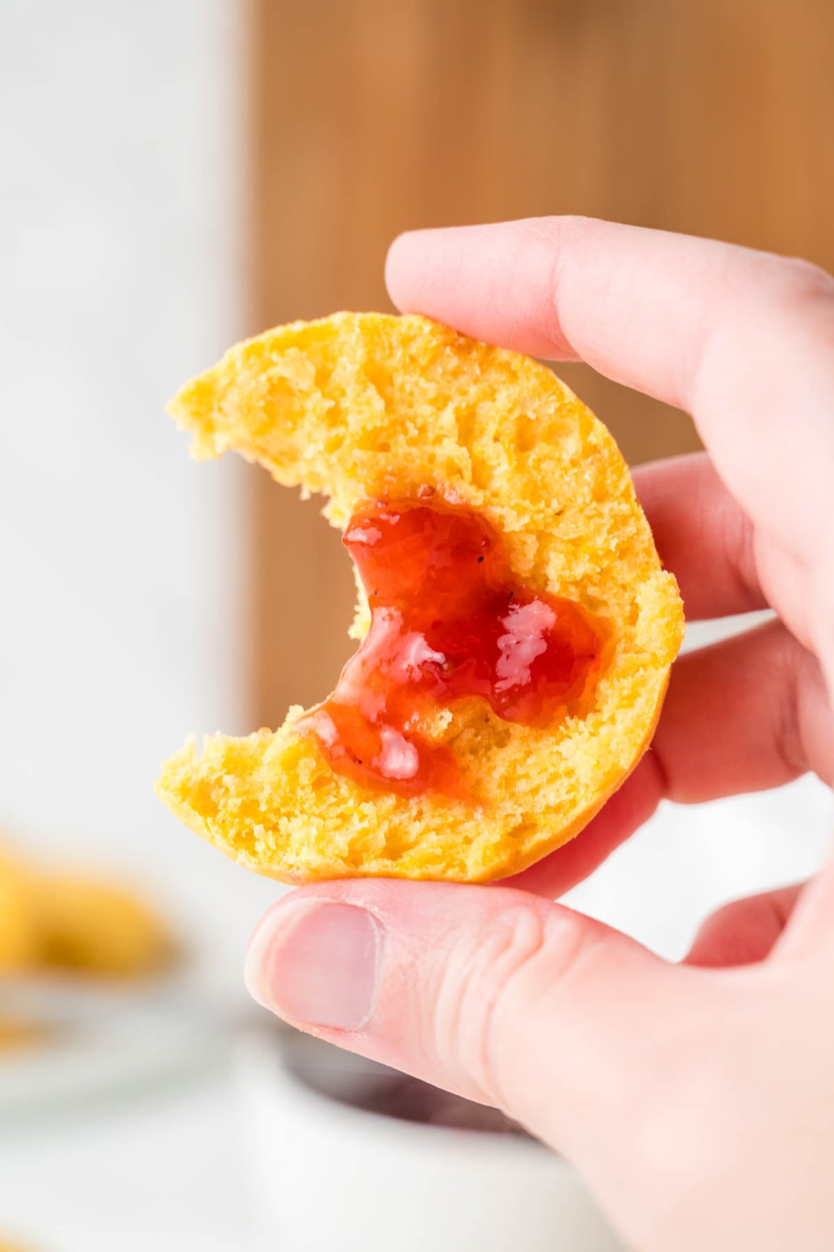 A hand holds a bitten piece of biscuit topped with a dollop of red jam.