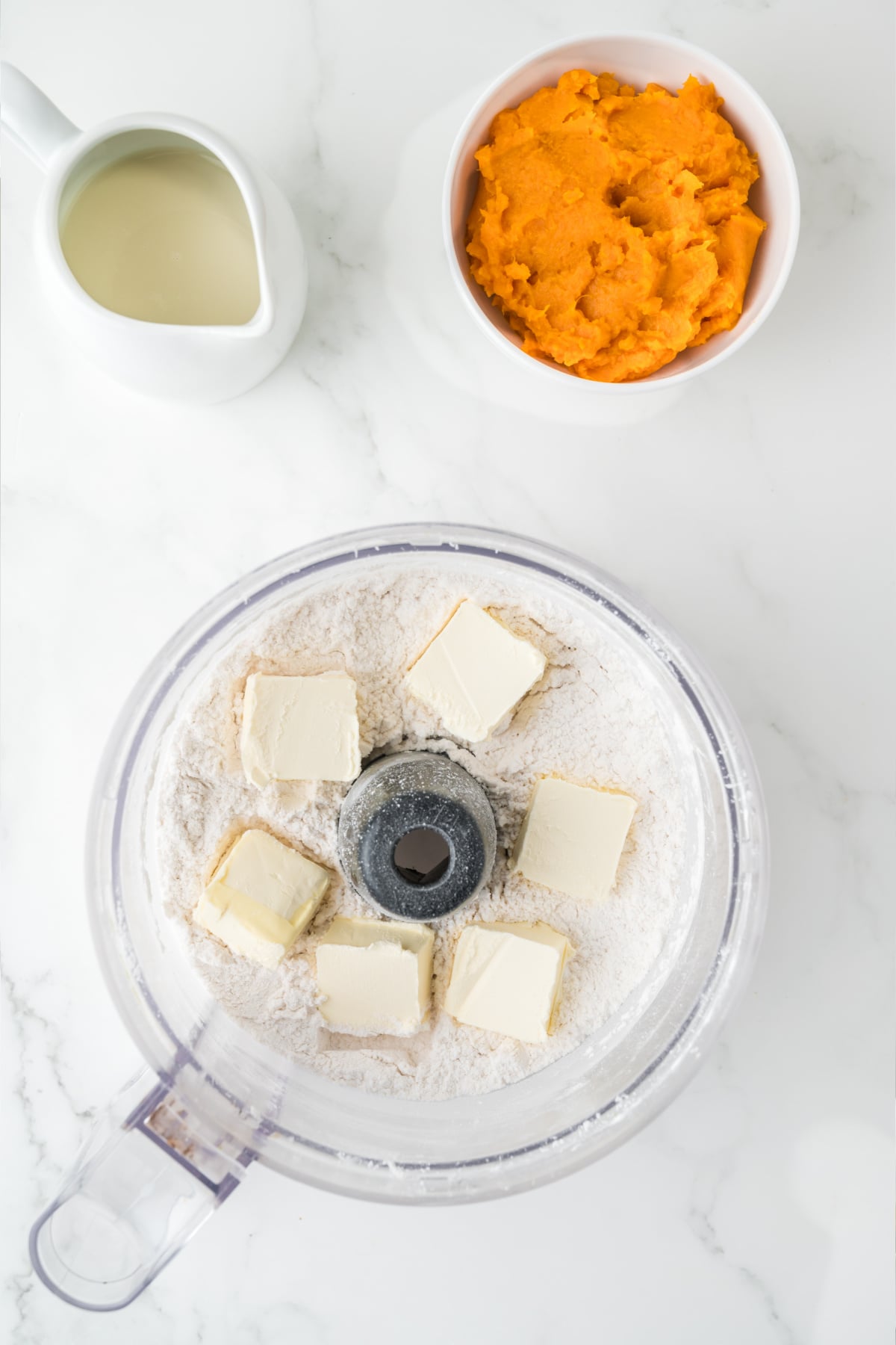 A food processor bowl containing flour and cubes of butter, with a bowl of mashed sweet potatoes and a small pitcher of milk nearby.