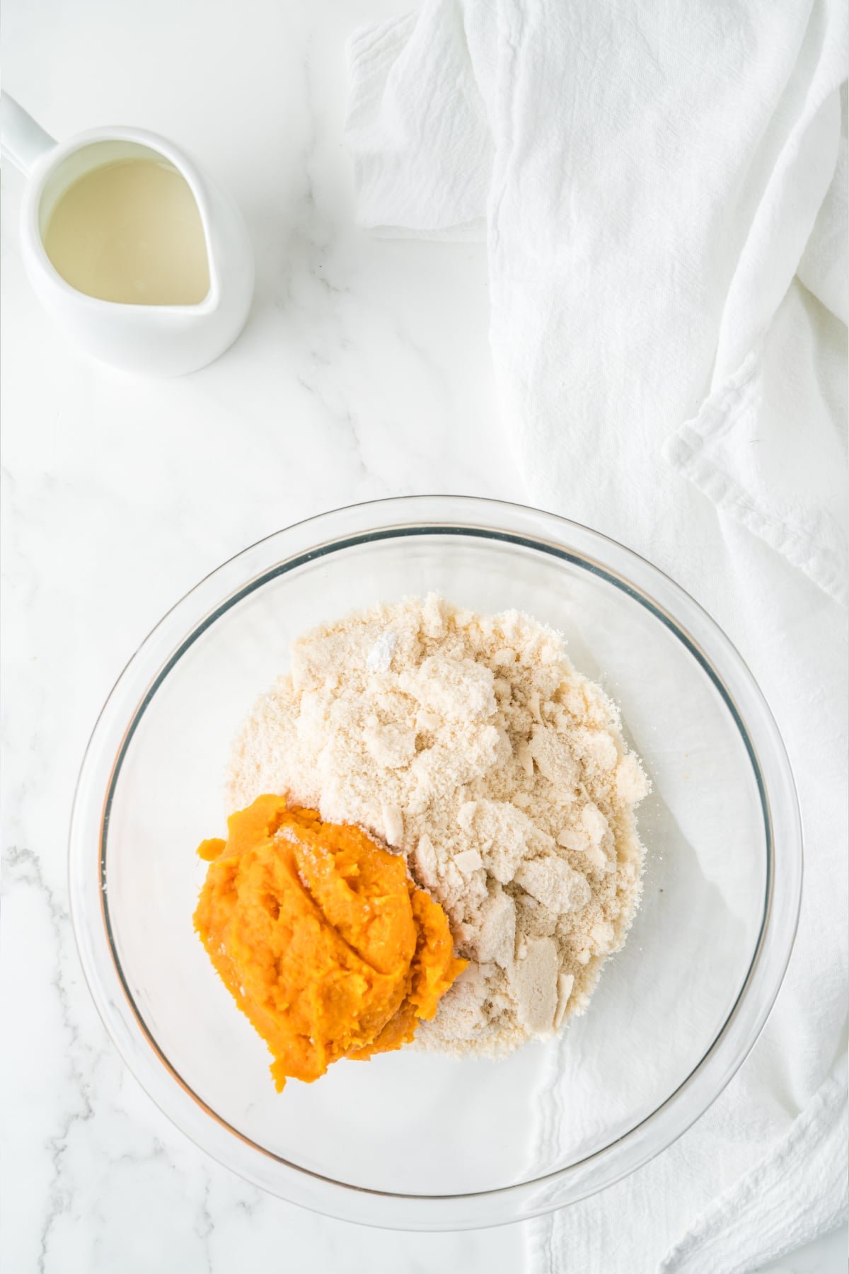 A glass bowl containing a mound of dry flour mixture next to a scoop of mashed sweet potatoes, with a small pitcher of milk nearby.