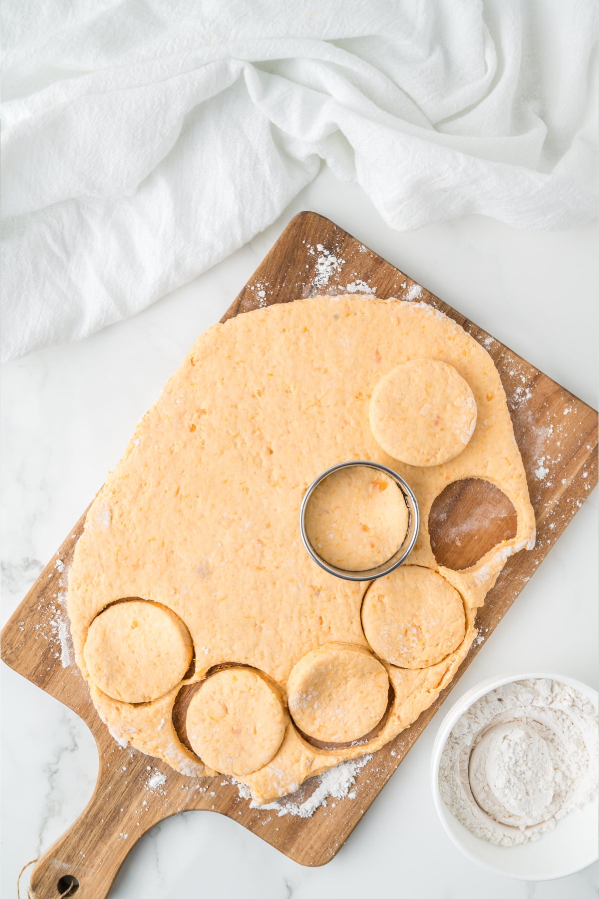 Dough rolled out on a wooden cutting board with a circular metal cutter forming biscuit shapes, next to a bowl of flour.