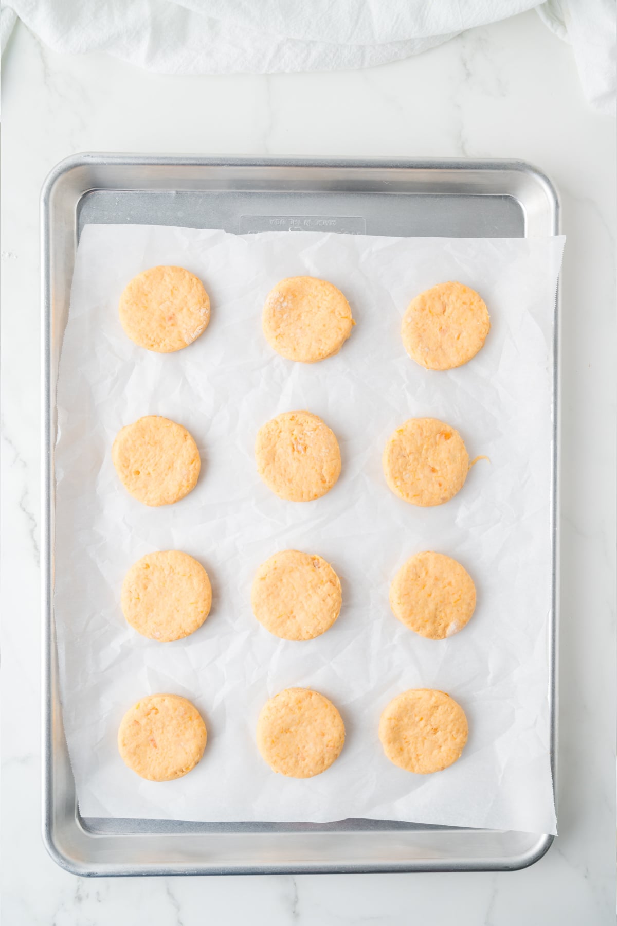 Twelve raw, circular dough portions arranged in a grid on a parchment-lined baking sheet.